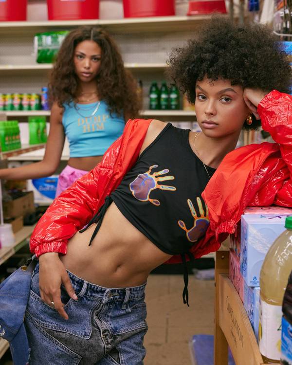 Two women in trendy clothing pose in a bodega. One woman wears a black crop top, jeans, and a red jacket; the other wears a blue tank top.