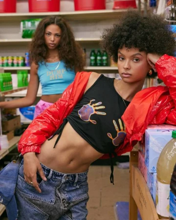 Two women in trendy clothing pose in a bodega. One woman wears a black crop top, jeans, and a red jacket; the other wears a blue tank top.