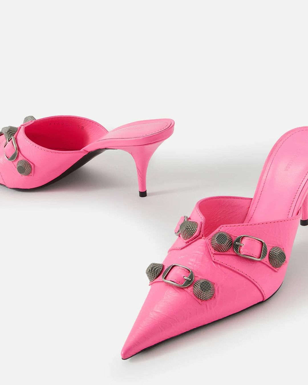 Pair of bright pink heeled mules with pointed toe, embellished with silver buckles and hardware on a plain white backdrop.
