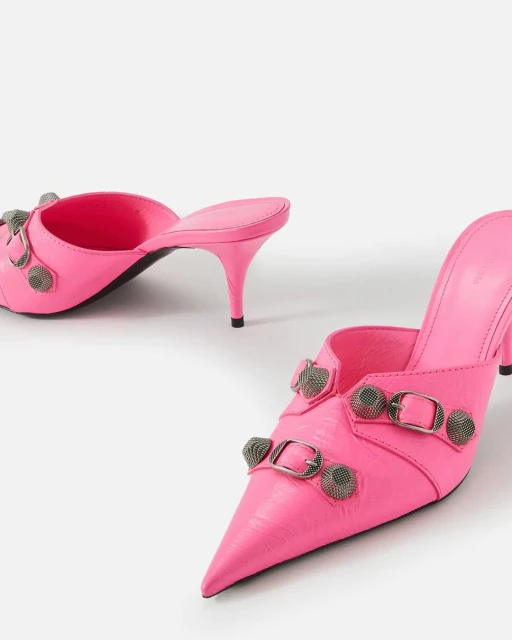 Pair of bright pink heeled mules with pointed toe, embellished with silver buckles and hardware on a plain white backdrop.