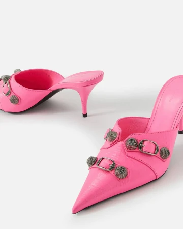 Pair of bright pink heeled mules with pointed toe, embellished with silver buckles and hardware on a plain white backdrop.