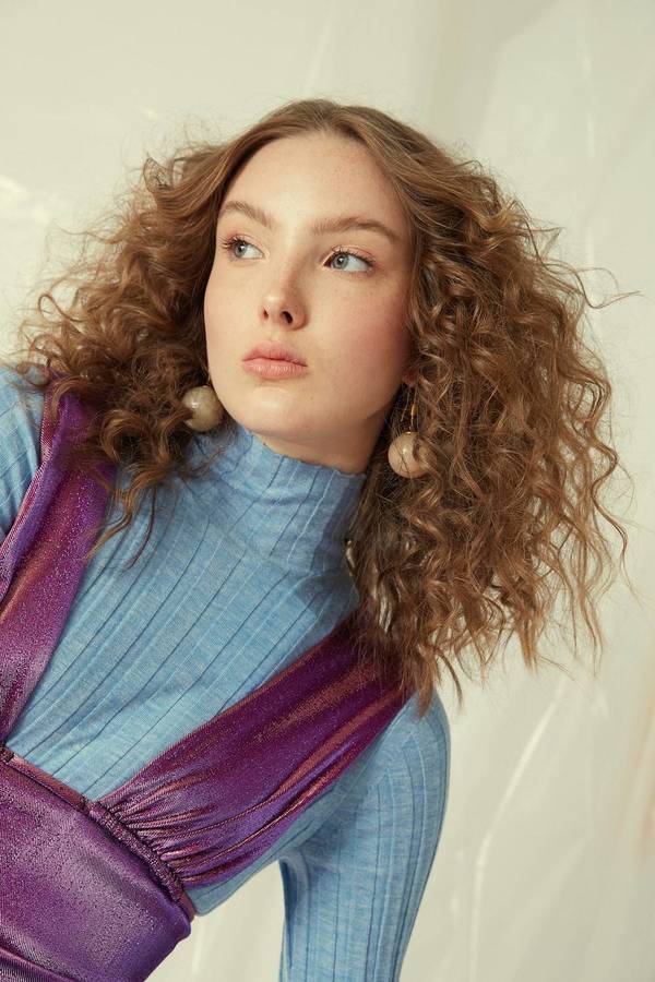 Woman with curly auburn hair, wearing a purple dress with a blue ribbed turtleneck, posing in a light-filled studio. Fashion shot.