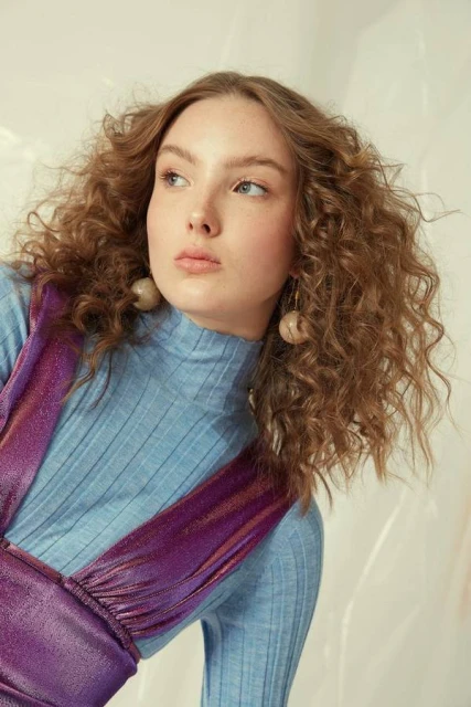 Woman with curly auburn hair, wearing a purple dress with a blue ribbed turtleneck, posing in a light-filled studio. Fashion shot.