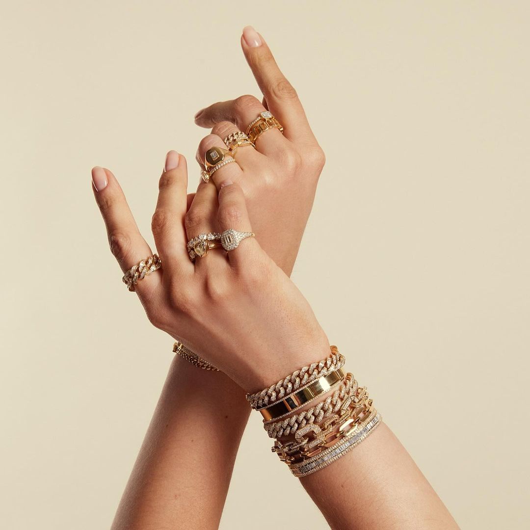 A person wearing gold jewelry including bracelets and rings against a beige background. Gold chain bracelets and rings with diamonds.