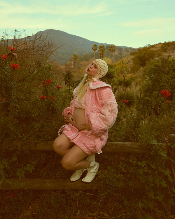 Pregnant woman in pink outfit poses outdoors. She's kneeling on a wooden fence amid greenery, hills, and a bright sky.