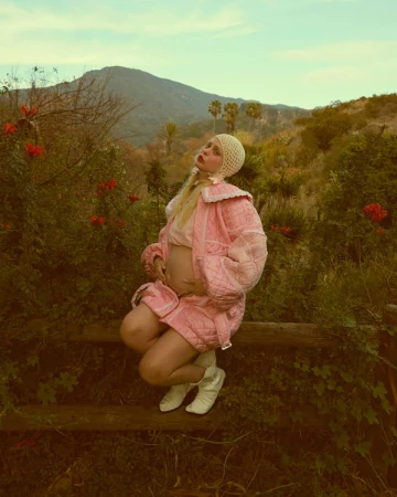 Pregnant woman in pink outfit poses outdoors. She's kneeling on a wooden fence amid greenery, hills, and a bright sky.