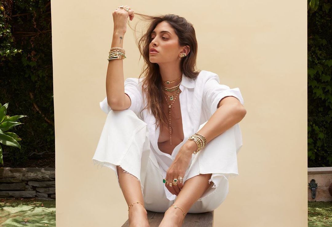 Woman in white shirt and pants posing with layered gold jewelry against a pale wall by photographer Katy Pritchett