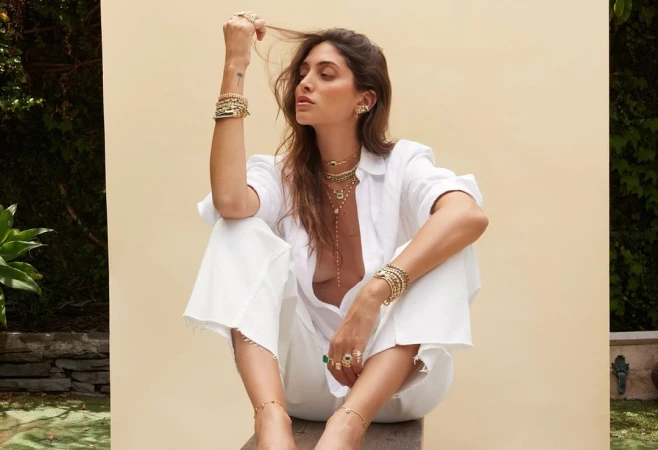 Woman in white shirt and pants posing with layered gold jewelry against a pale wall by photographer Katy Pritchett
