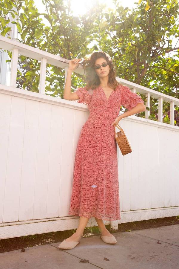 Woman in red patterned midi dress and sunglasses leaning against a white wooden fence under lemon trees