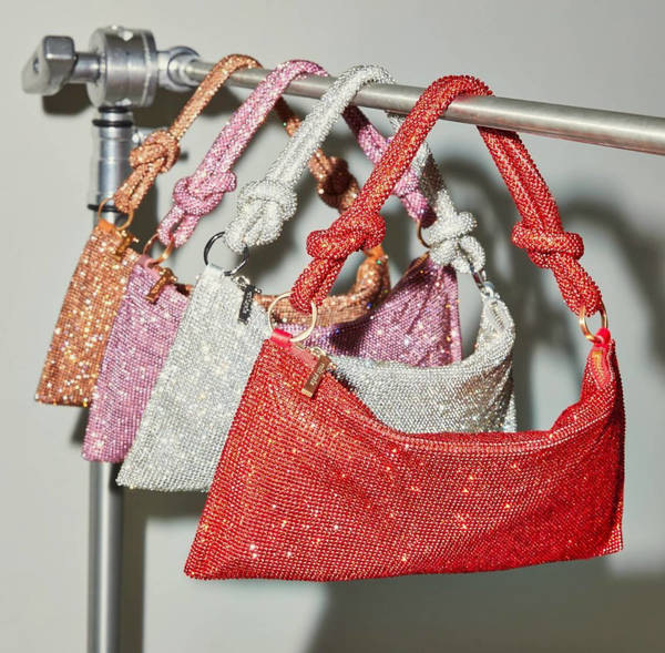 Four sparkling baguette bags in red, silver, pink, and bronze, with knotted handles, hang on a metal bar against a gray backdrop.