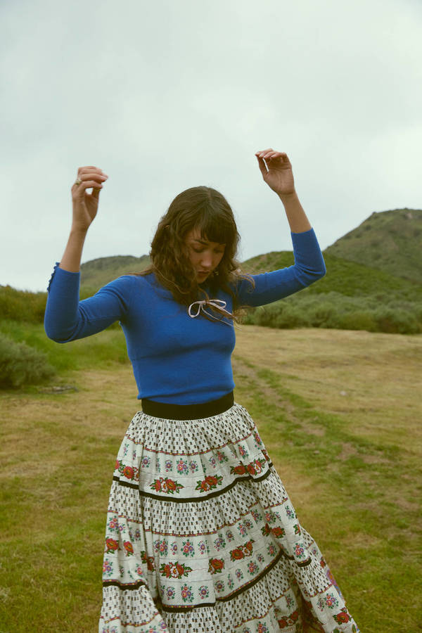 A woman in a blue top and patterned skirt dances on a grassy hill under an overcast sky.