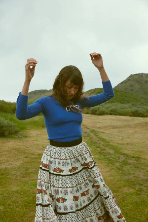 A woman in a blue top and patterned skirt dances on a grassy hill under an overcast sky.