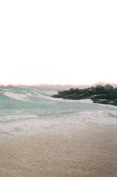 Sandy beach with crashing ocean waves meeting dark rocky shoreline under bright sky