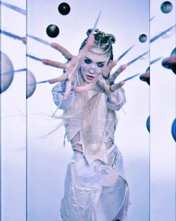 Pale aesthetic image of a woman with fantasy makeup and braided hair, posing with large decorative nails in front of a sci-fi set.