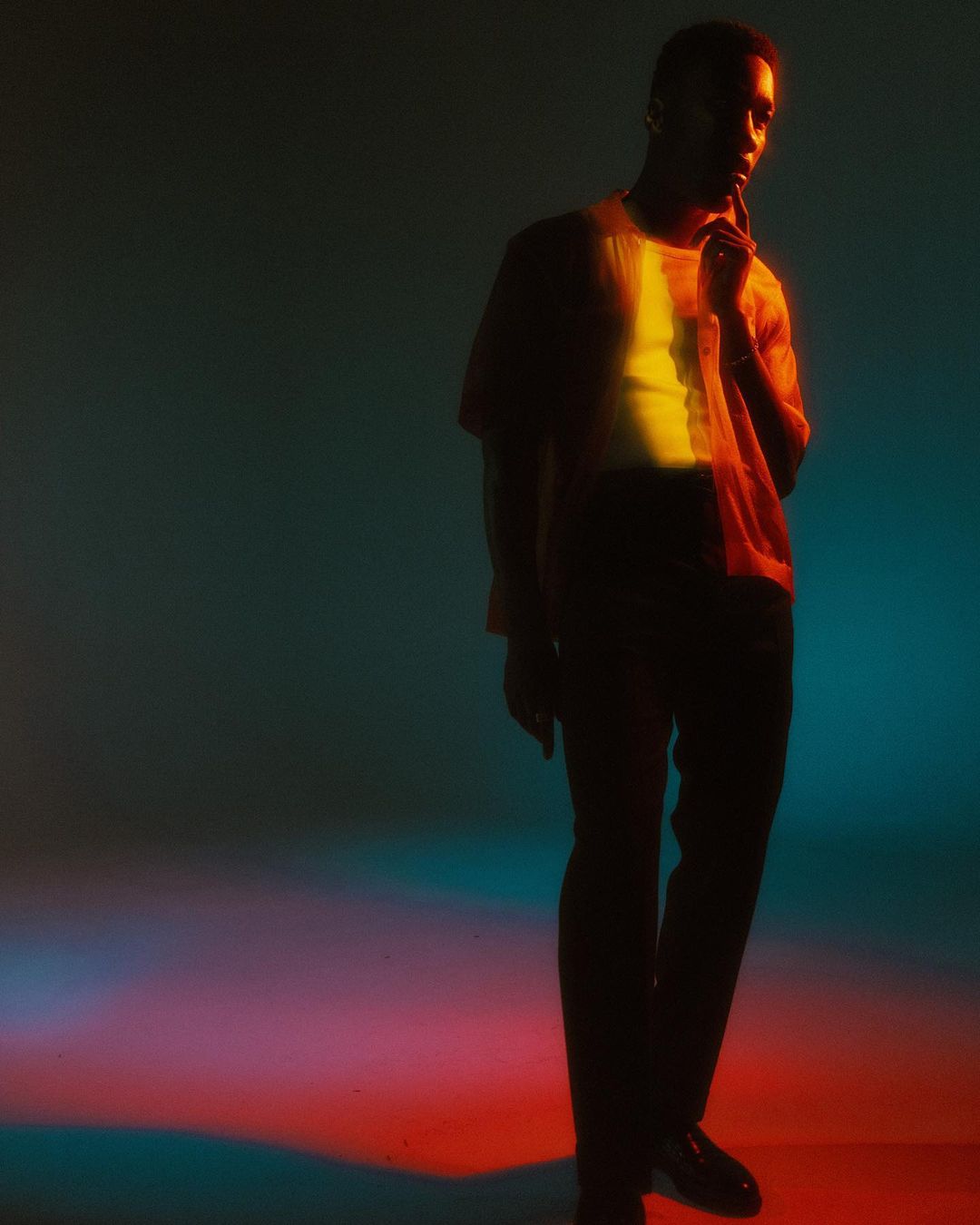 Portrait of a black man in a casual shirt and dark pants in a studio setting with dynamic red, yellow, and teal lighting.