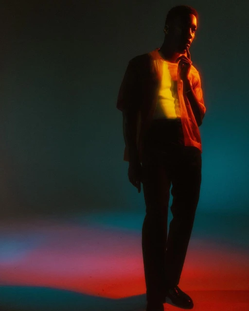 Portrait of a black man in a casual shirt and dark pants in a studio setting with dynamic red, yellow, and teal lighting.