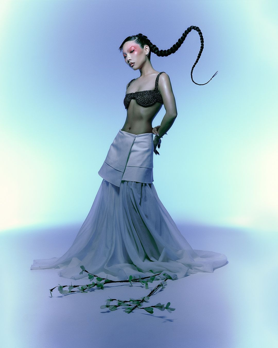 Model with a long braided ponytail wearing a dark beaded bra and white tiered skirt against a blue gradient backdrop.
