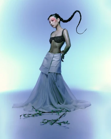 Model with a long braided ponytail wearing a dark beaded bra and white tiered skirt against a blue gradient backdrop.