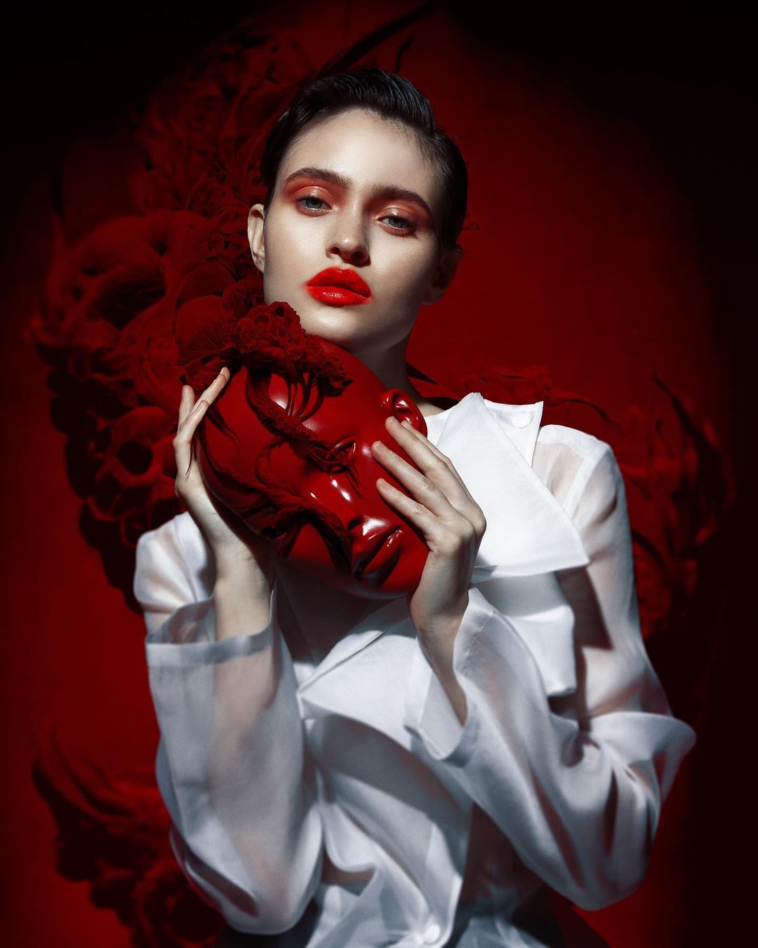 Portrait of a model holding a shiny red mask, wearing a white blouse against a dark red background with smoke effects.