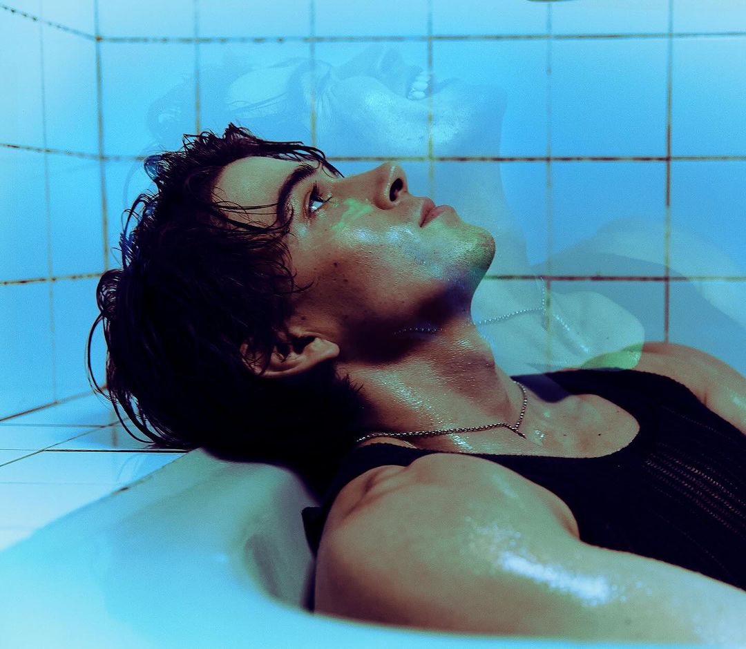 Man lying in a bathtub with blue lighting. He is wearing a black tank top and a silver necklace. Wet hair and wet skin.
