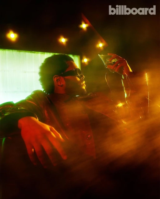 Portrait of The Weeknd holding a cocktail glass in moody, warm lighting. Billboard logo visible in the top right corner.