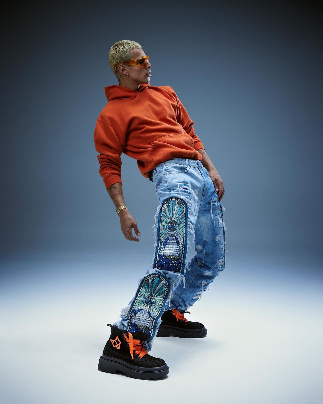Full shot of adult male in an orange hoodie and jeans with graphic art. He is wearing sunglasses and black shoes with orange laces.