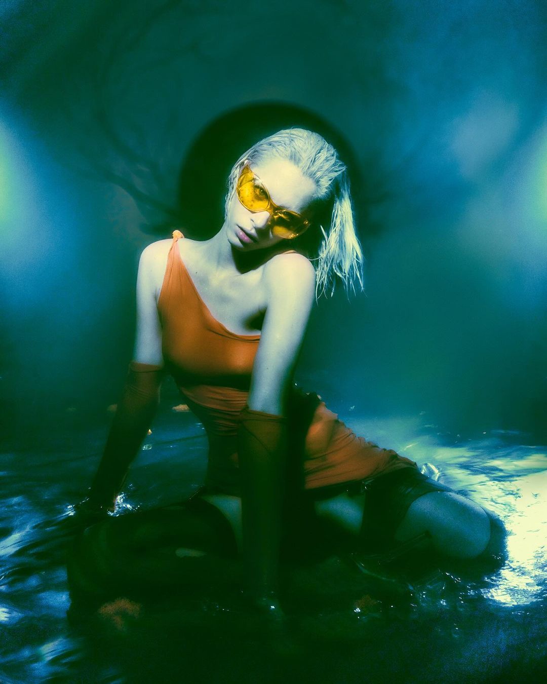 Blonde woman in orange dress and yellow glasses kneels in shallow water with a moody, artistic, and slightly surreal aesthetic.