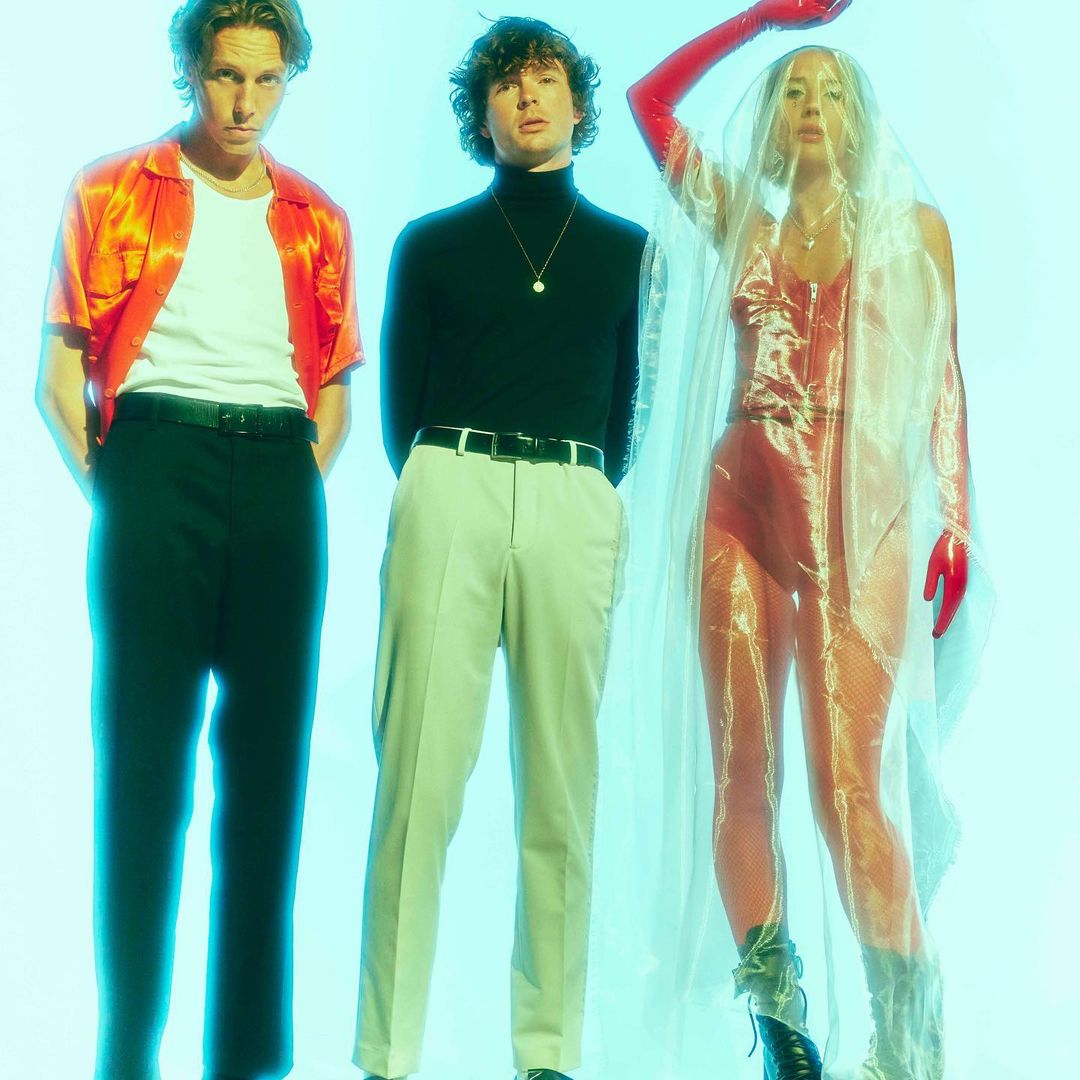 Three people pose in a studio: two men in shirts and pants, and one woman in a red bodysuit, gloves, and a transparent fabric.