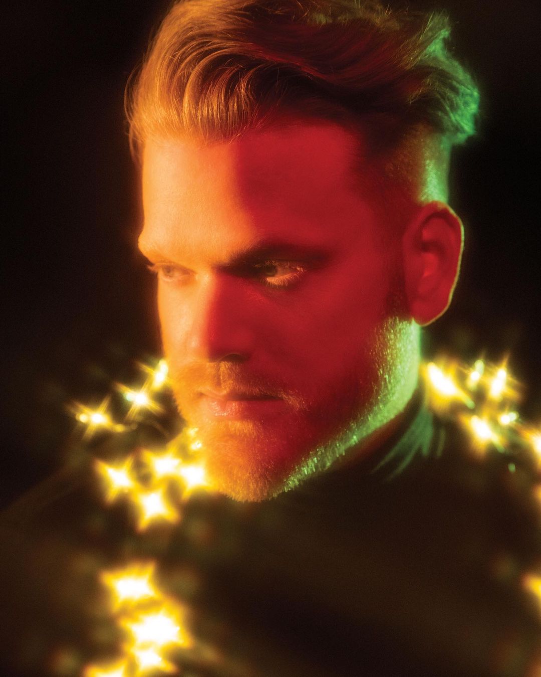 Portrait of a red-haired adult man with fair skin, posing indoors with golden star-shaped lights around his neck against a dark background.