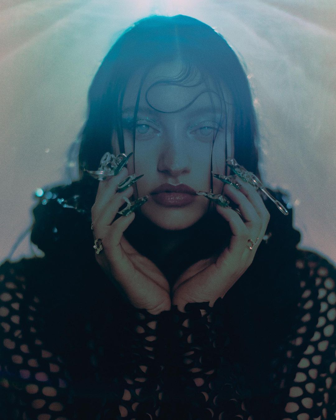 Close-up studio portrait of a woman with long dark hair, light skin, and long decorated fingernails, wearing a fishnet-style top, illuminated by blue light.