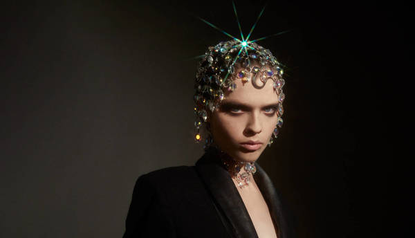 A person with decorative jeweled headwear, in dark studio lighting with a low angle, wearing a black suit jacket and jeweled choker.