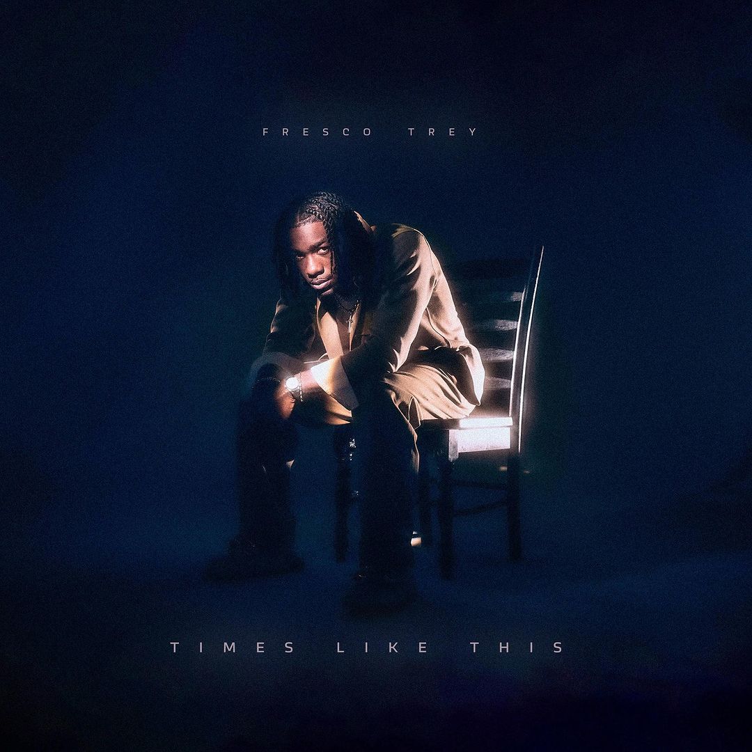 Darkly lit album cover featuring a young Black male artist seated on a wooden chair against a dark blue gradient background.