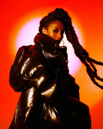 Portrait of a woman wearing a glossy black puffer jacket against a striking orange and red backlit background by Brian Ziff.