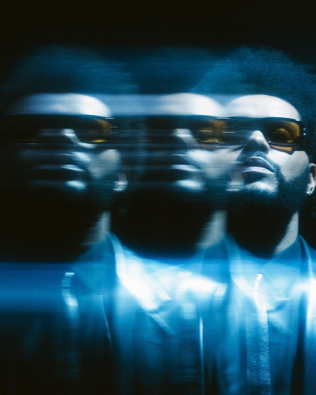 Blurred image of an adult male with dark hair and a beard, wearing sunglasses and a button-down shirt, set against a dark background.