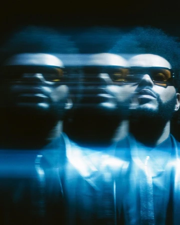 Blurred image of an adult male with dark hair and a beard, wearing sunglasses and a button-down shirt, set against a dark background.