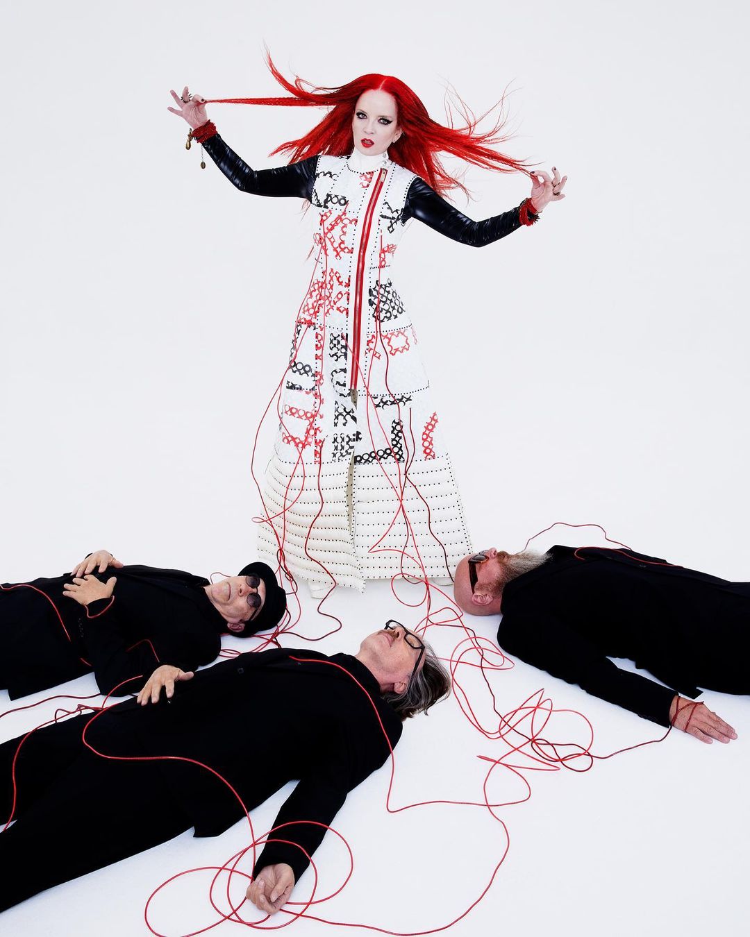 Artistic portrait of a woman with red hair standing over three men lying on the floor connected by a red string in a studio.