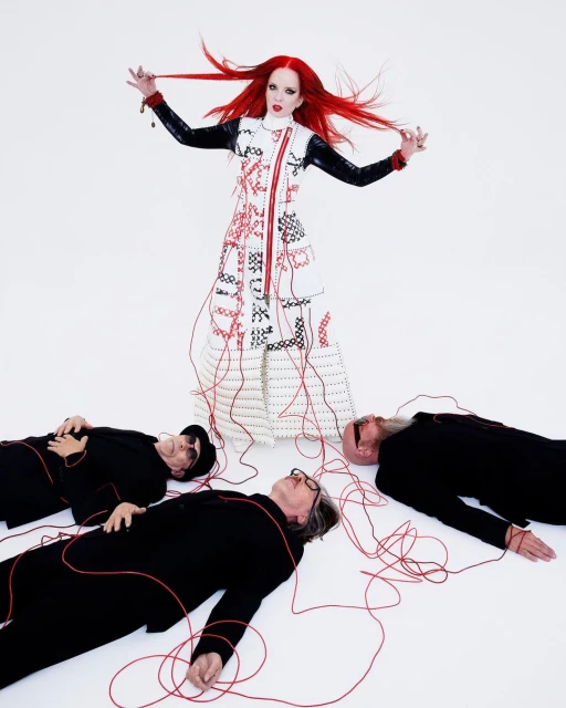 Artistic portrait of a woman with red hair standing over three men lying on the floor connected by a red string in a studio.