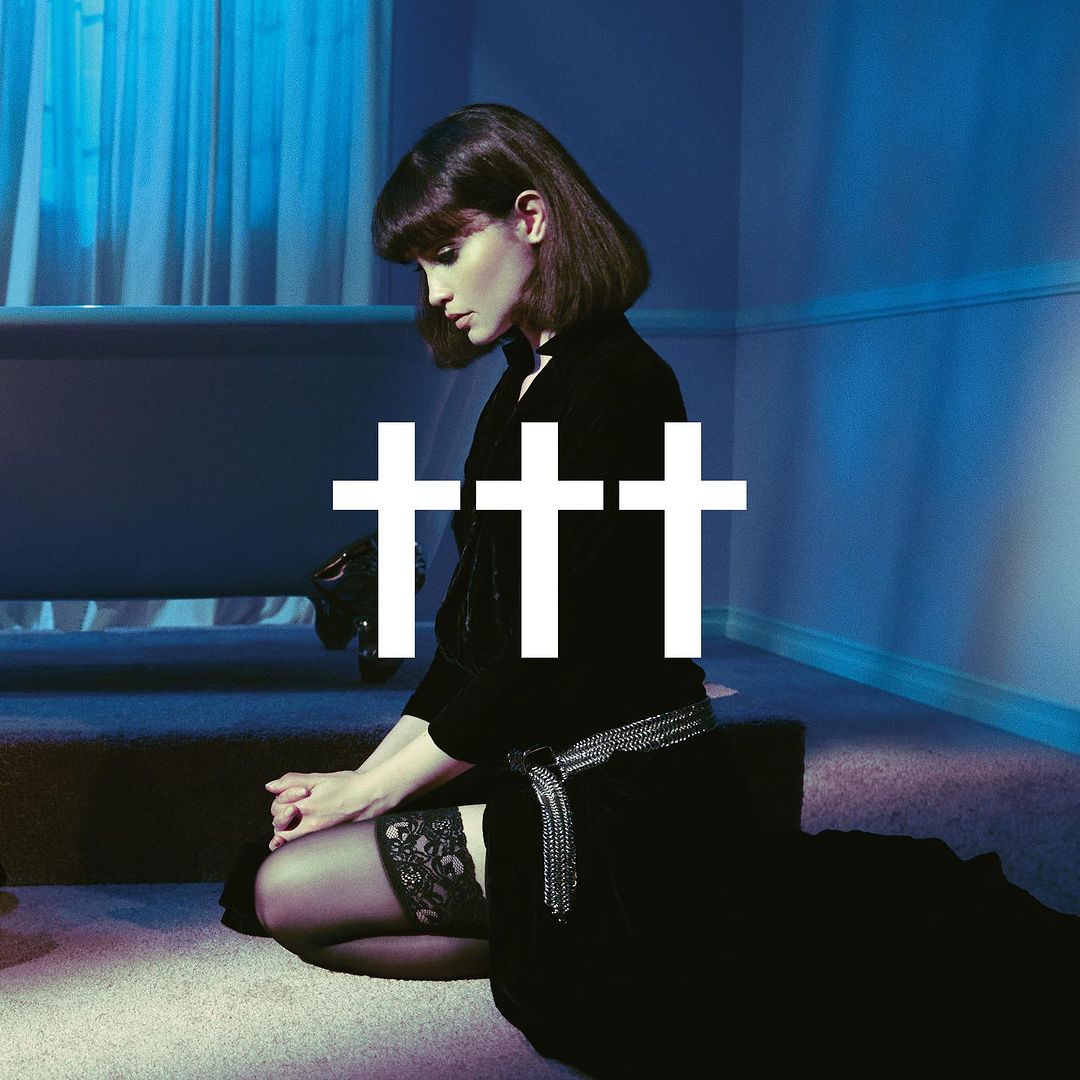 A brunette adult woman kneels in a black dress with white crosses, inside an indoor space with blue walls and carpeted floors.