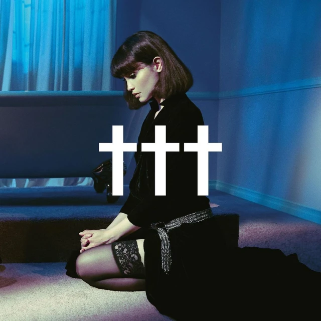 A brunette adult woman kneels in a black dress with white crosses, inside an indoor space with blue walls and carpeted floors.