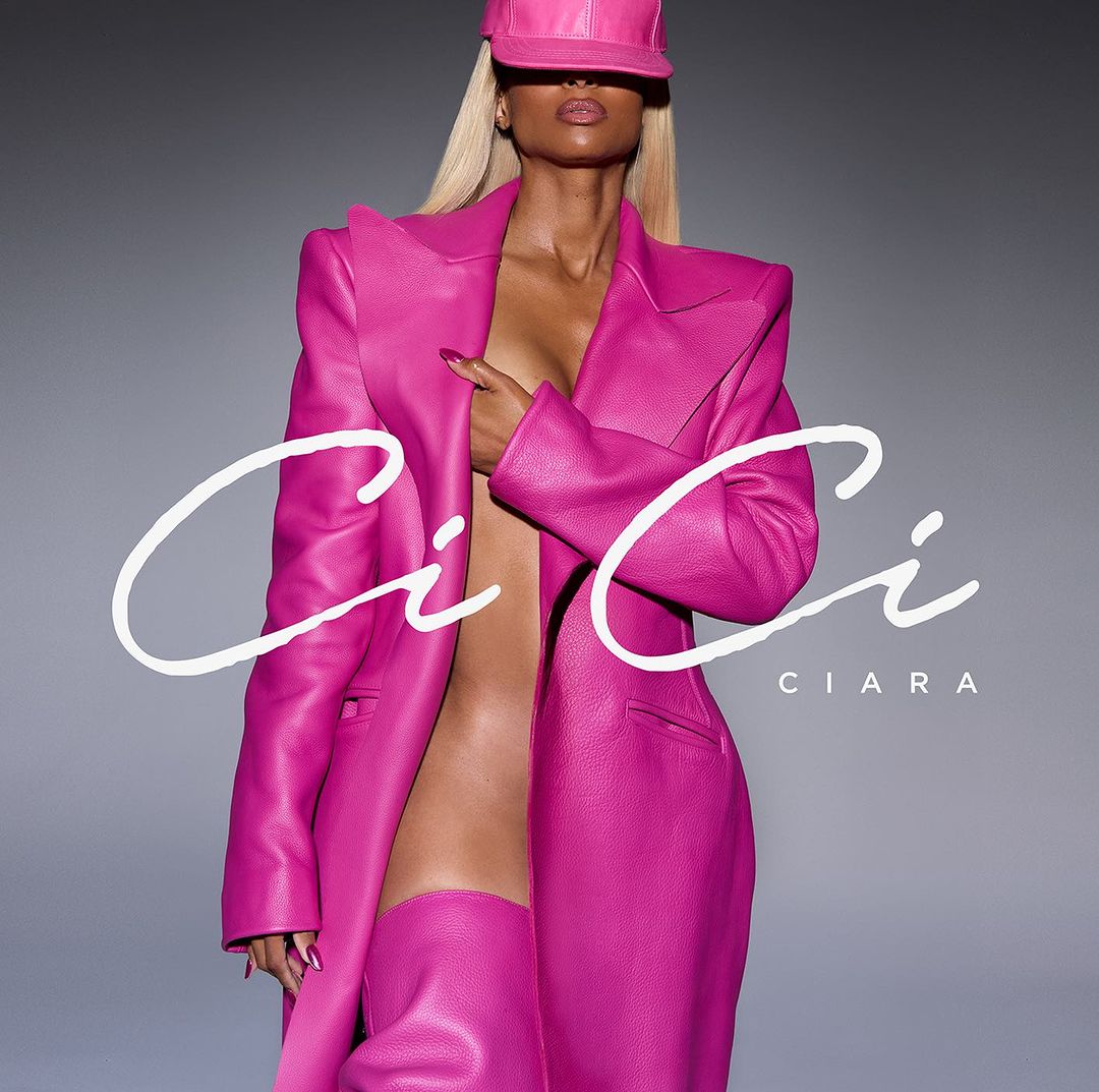 Singer Ciara poses wearing a pink leather outfit consisting of a hat, jacket, and thigh-high boots against a gray background.
