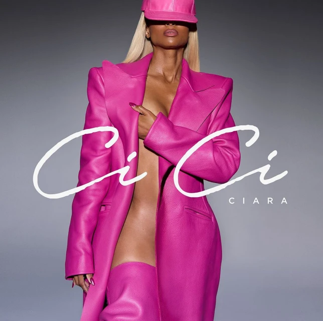 Singer Ciara poses wearing a pink leather outfit consisting of a hat, jacket, and thigh-high boots against a gray background.