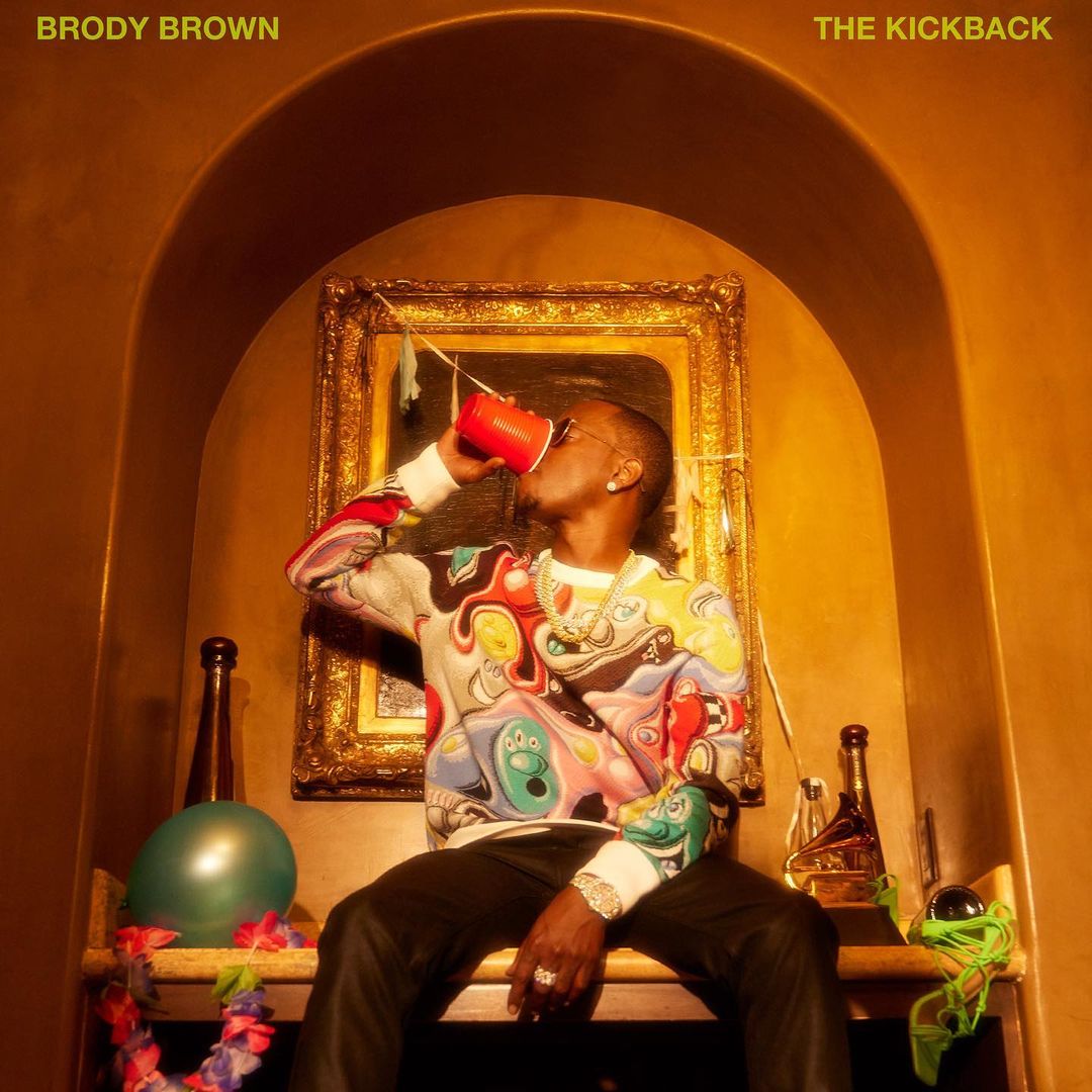 Adult male wearing sunglasses and colorful sweater seated against a gold background, drinking from a red cup, with bottles and decorations.