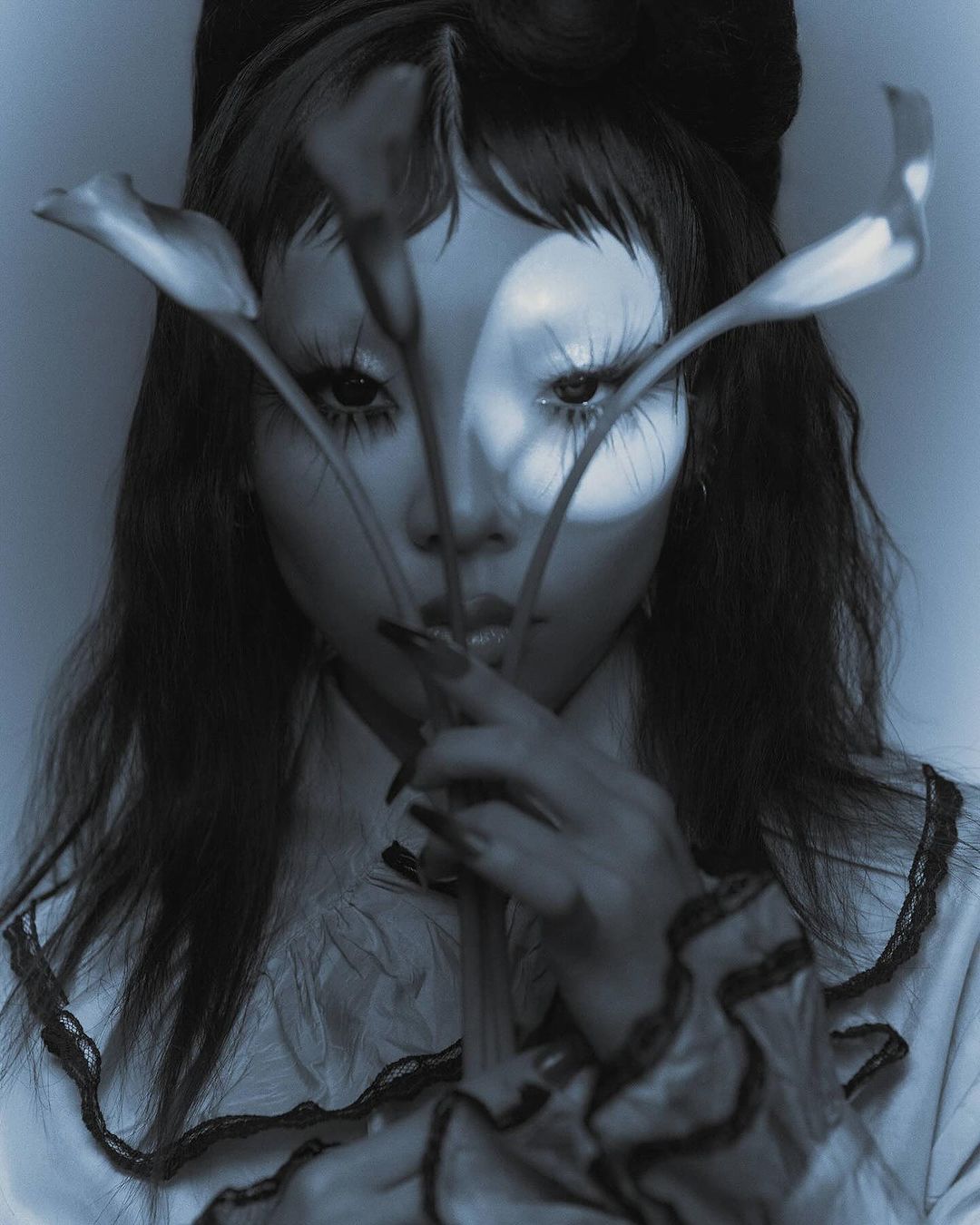 Monochromatic close-up portrait of a woman with striking makeup holding calla lilies in front of her face in an indoor setting.