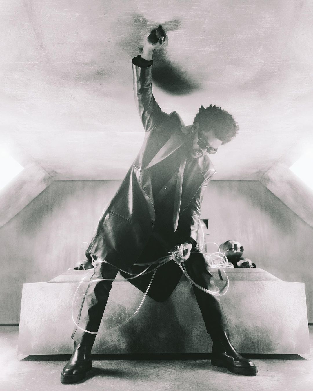 Monochrome portrait of a person in a black leather trench coat, sunglasses, and boots, standing in a room with a raised platform.