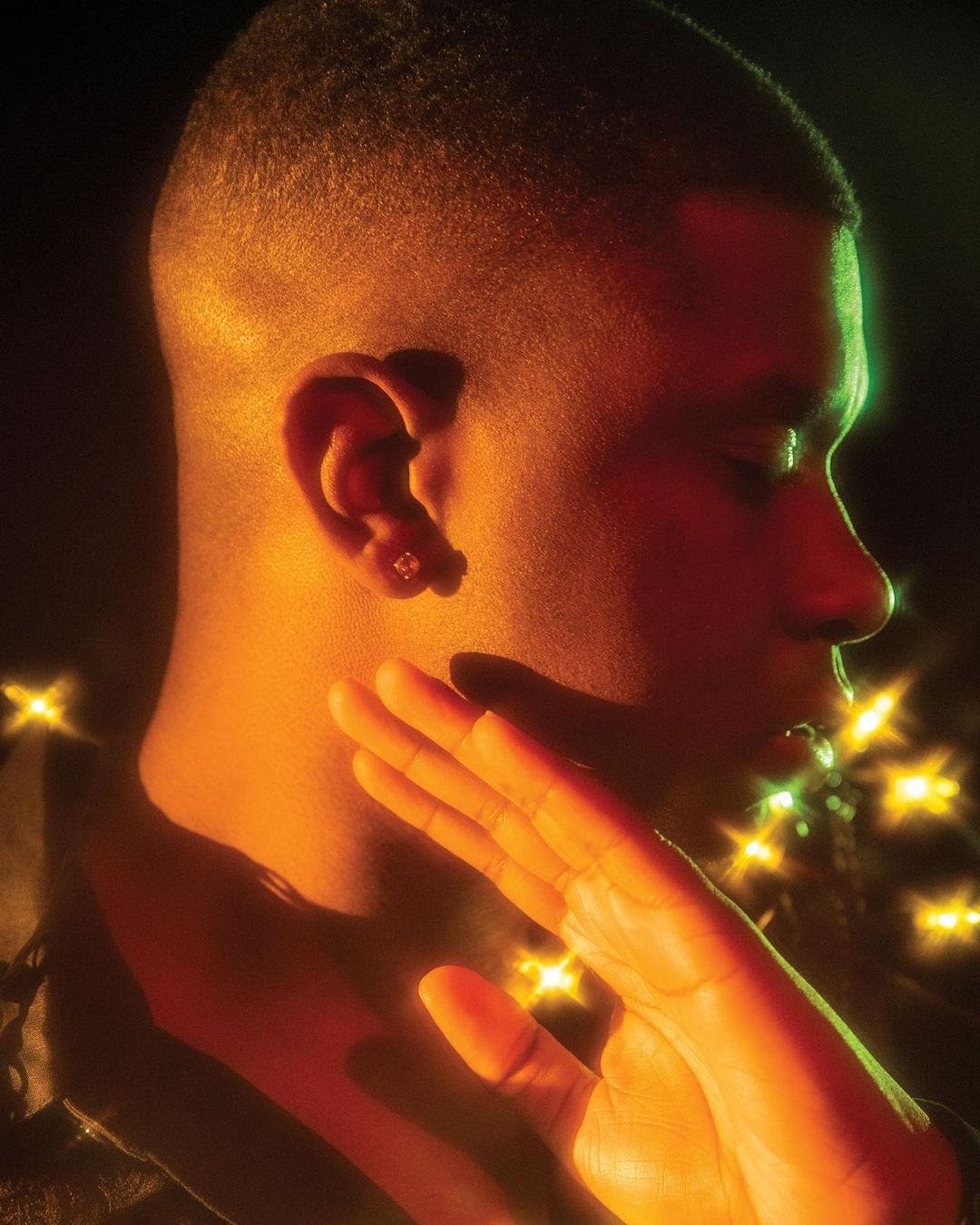Close-up portrait of a young adult male with a short haircut and a diamond stud earring, posing in orange and green lighting.