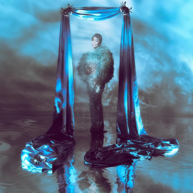 Woman in an elaborate dark gown posing beneath a draped archway against a dreamlike sky in a studio shot with digital effects.