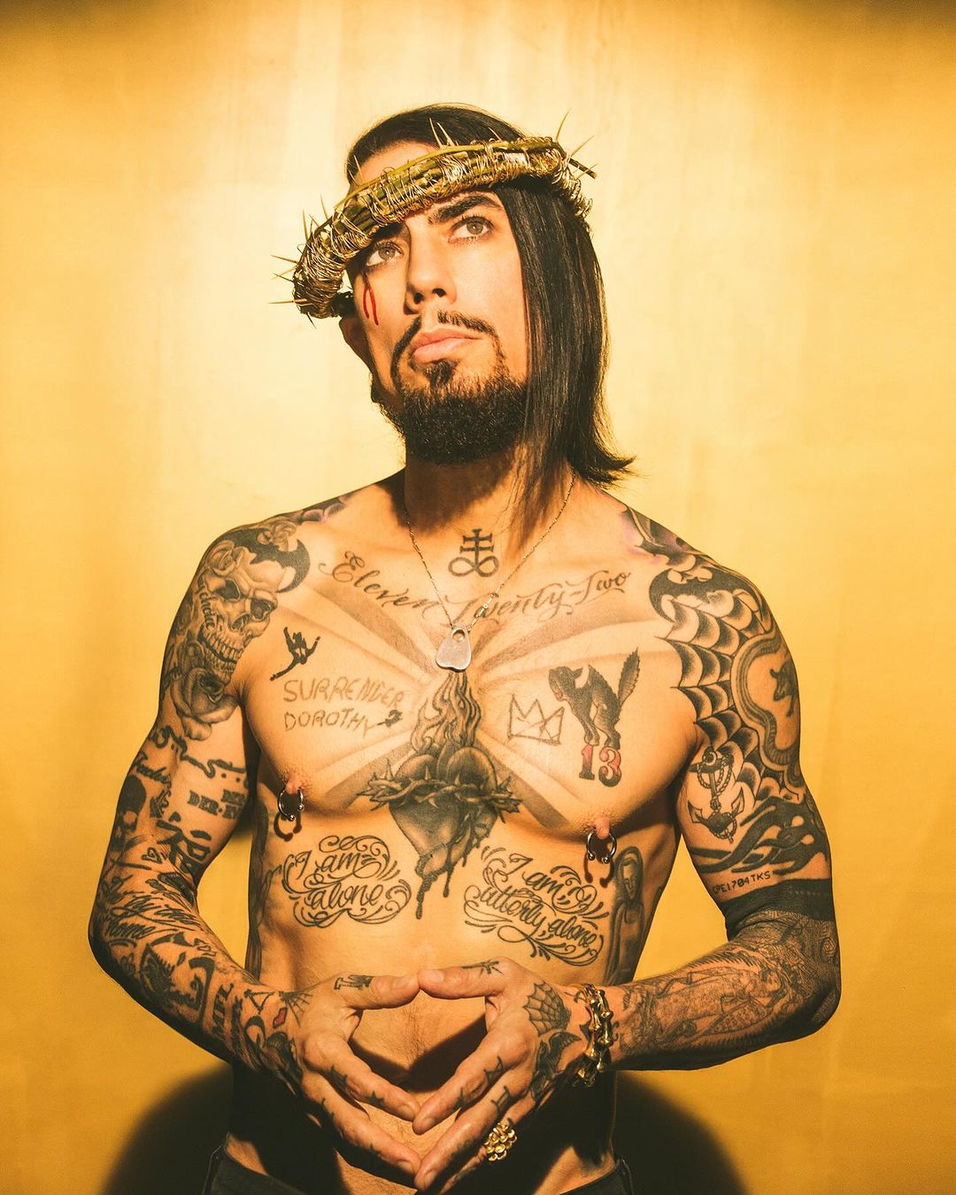 Tattooed adult male with a crown of thorns on head, posing against a warm yellow backdrop. Heavily tattooed, shirtless, indoor portrait.