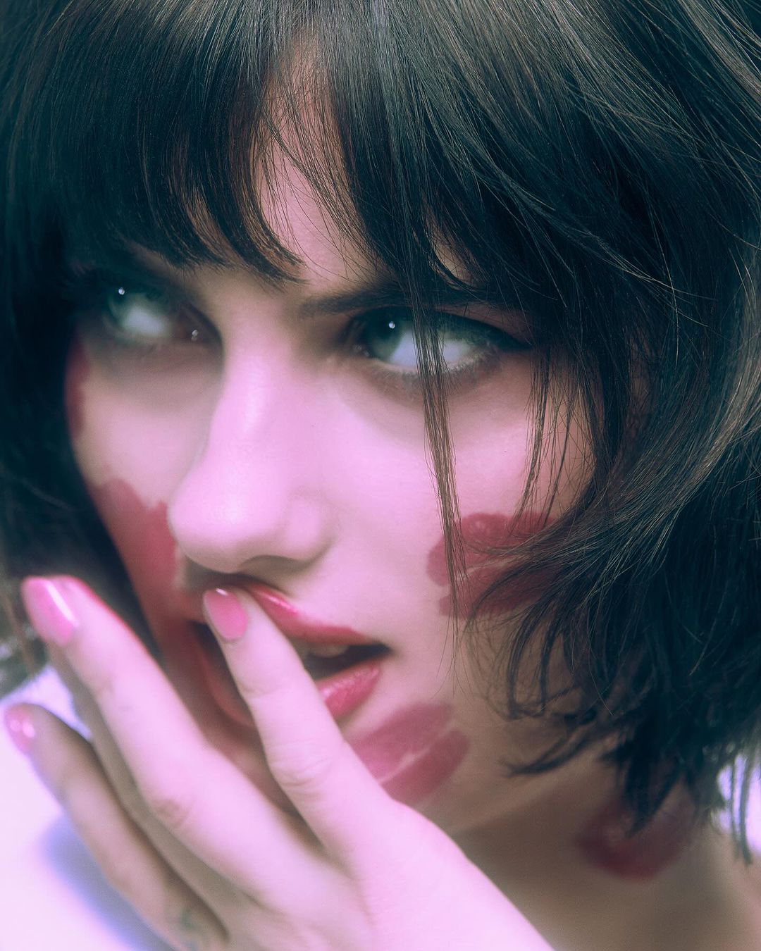 Close-up portrait of a young woman with lipstick marks on her face and hand touching her lips in a beauty-themed shot.