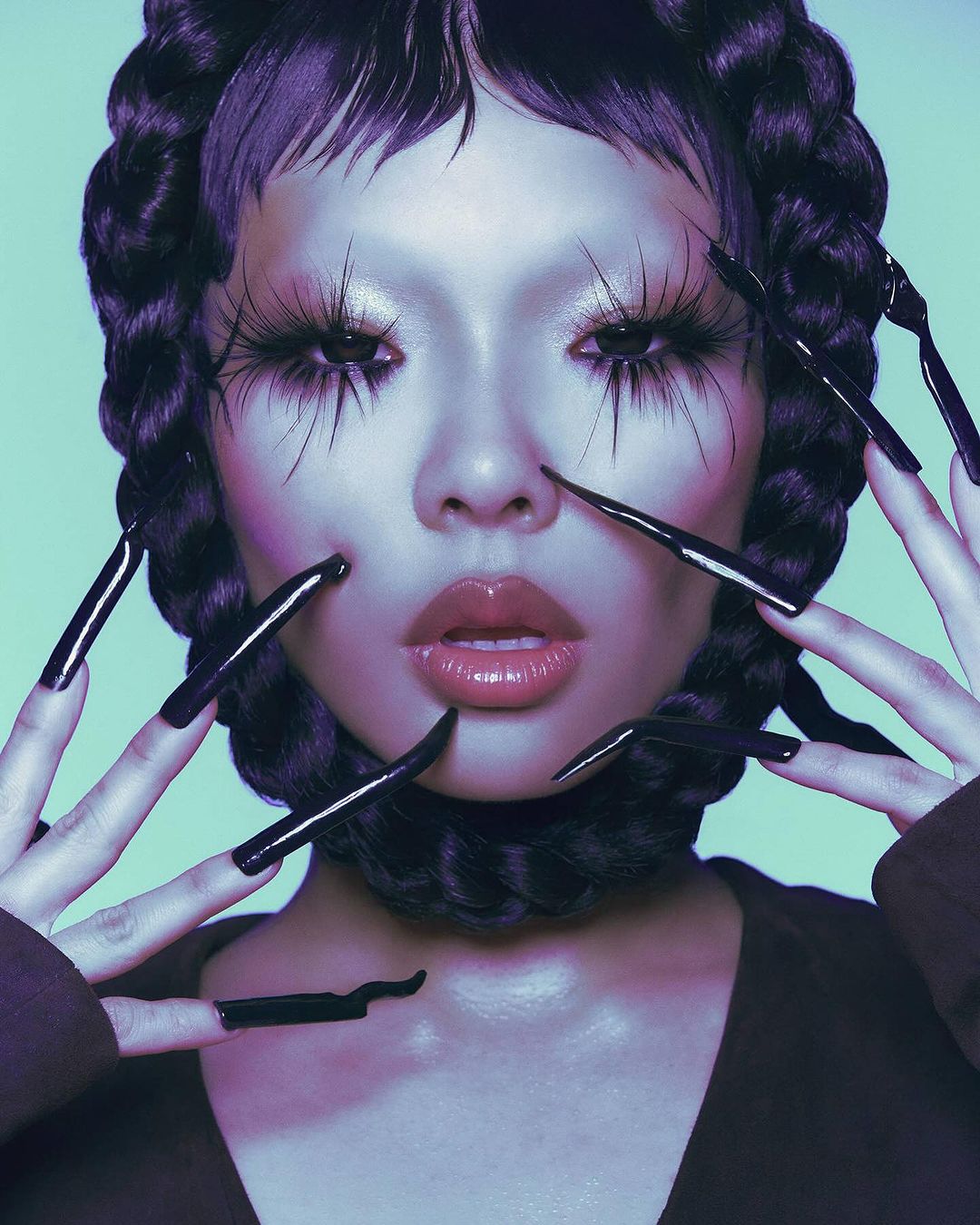Close-up portrait of a woman with extravagant eyelashes and glossy lips, posing with black stiletto nails against a light-blue backdrop.