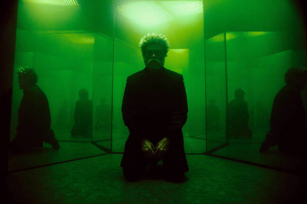 A man kneeling in a mirrored chamber illuminated by vibrant green light, creating multiple reflections.